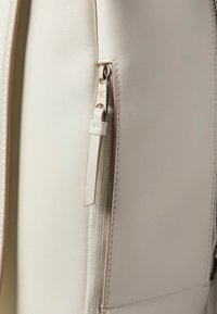 Detail bílého koženého kabelky s hlavním zipem s kovovým jezdcem a prošitými okraji. Texturovaný látkový akcent blízko zipu.