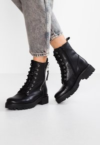 Bottes chevilles en cuir noir avec lacets, fermeture éclair latérale, semelle épaisse et dessus texturé. Accents bicolores sur les lacets et la fermeture éclair.