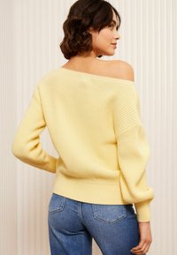 Pulover galben tricotat, cu umeri goi, cu textură cu nervuri, mâneci lungi și o croială lejeră, combinat cu blugi de denim de culoare albastru mediu.