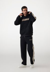 Čierna súprava s kapucňou a teplákmi s béžovými bočnými prúžkami; obsahuje prednú vrecko, rebrované manžety a veľké logo „GUESS“ v zlatej farbe na oboch kusoch.