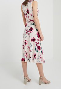 Blumenkleid ohne Ärmel in Creme mit pinken und dunkelroten Mustern. A-Linien-Form, fließender Stoff, taillierter Schnitt. Ergänzt durch beiger hohe Sandalen.