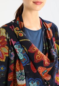 Kvinna som bär en mörk marinblå sjal med stora färgglada blommor och fjärils mönster över en blå skjorta, håret uppsatt i en hästsvans.