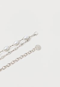Brățară din lanț argintiu cu perle rotunde albe false și accente clare din cristal, fixată cu un cârlig lobster și detaliu cu etichetă de logo.
