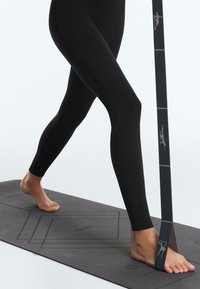 Osoba v černých legínách se protahuje naboso na tmavé jógové podložce s odporovou gumou upevněnou kolem jedné nohy.