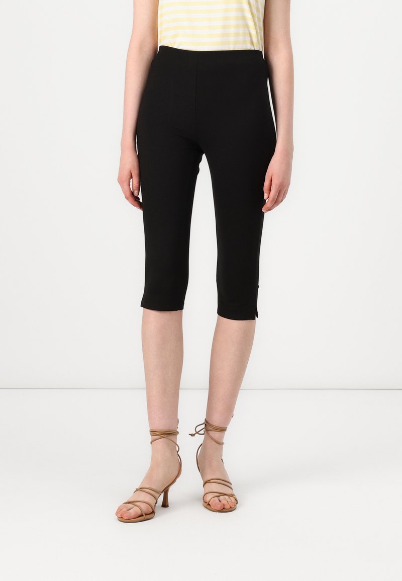 Svarta capri leggings har en slät elastisk midja och en kort änd, kombinerat med tan färgade remsandaler med hög klack. Bakgrunden är neutral vit.