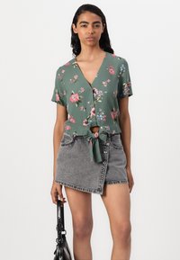 Camicia verde a maniche corte con motivo floreale, scollo a V e orlo annodato, abbinata a una minigonna in denim grigia con chiusura frontale a bottoni.
