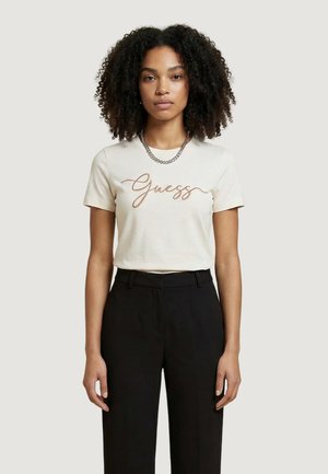 Mujer con cabello rizado que lleva una camiseta blanca de manga corta con la palabra "Guess" en letras cursivas, combinada con pantalones negros de tiro alto y un collar de cadena plateada.