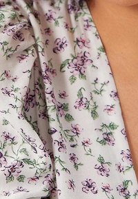 Vitt tyg med små lila blommor och gröna blad, delvis draperat över en persons axel och övre bröst.