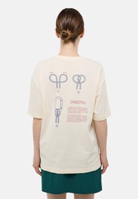 T-shirt en coton beige à manches courtes, avec des illustrations de noeuds en corde et un texte au dos en imprimé bleu et rouge.