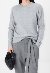 Grau gestrickter Pullover mit geripptem Kragen und Bündchen, kombiniert mit grauen hoch taillierten Hosen mit einem gebundenen Gürtel. Hält eine schwarze Ledertasche.