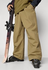 Snøbrettbukser i khaki-stoff, løs passform, med flere lommer og forsterkede sømmer, kombinert med svarte skistøvler og en ski.