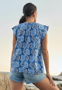Blue patterned top featuring white shell motifs, short flutter sleeves, and a relaxed fit, paired with light denim shorts.