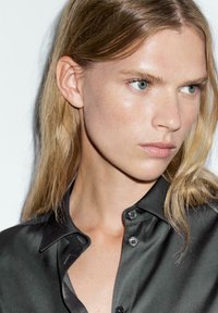 Camicia nera in satin con bottoni e colletto strutturato, caratterizzata da una texture liscia e una vestibilità morbida. Il modello ha capelli lunghi e chiari e trucco naturale.