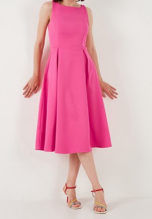 Roze mouwloze jurk met een getailleerde bodice, een uitlopende rok en plooien. Het model heeft het gecombineerd met meerkleurige sandaaltjes met bandjes.