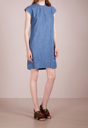 Žena v modrých džínových šatech bez rukávů s předními švovými detaily, doplněná hnědými sandály na vysokém podpatku s třásněmi, stojící na béžové podlaze.