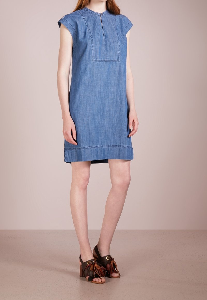 Femme portant une robe en denim bleu sans manches avec des détails de couture sur le devant, associée à des sandales à talons hauts marron avec des pompons, debout sur un sol beige.