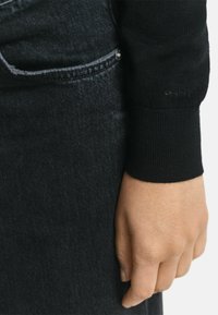 Schwarzer gerippter Pullover-Manschette kombiniert mit dunkler Jeans, die eine glatte Textur und dezente Nähte zeigt. Die Marke "GANT" ist auf der Manschette geprägt.