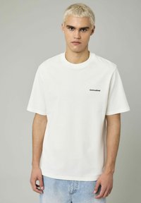 T-shirt en coton blanc avec des manches courtes et un col ras du cou. L'avant présente une broderie verte avec le mot "Untroubled". Coupe décontractée avec une texture lisse.