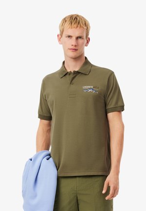 Joven vestido con una camiseta polo de Lacoste de color verde oliva con un logo de montaña, sosteniendo una sudadera plegada de color azul claro en una mano.