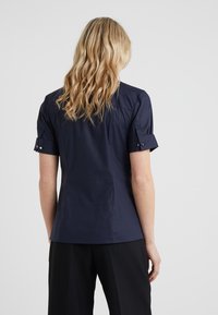 Donna bionda che indossa una camicia blu navy a maniche corte con risvolti e pantaloni neri, in piedi con la schiena rivolta contro uno sfondo bianco.
