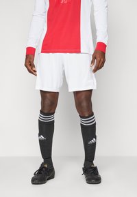 Balts futbola krekls ar sarkanu dizainu, balti šorti, melnas zeķes ar trim baltām svītrām, un melni futbola apavi ar teksturētu virsmu.