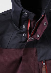 Close-up van de donkere bordeauxrode en zwarte jas kraag met een rode rits en zilveren drukknoopjes die gedeeltelijk vastgemaakt zijn.