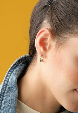 Gold hoop earring featuring a colorful gemstone design. Worn on the ear with a denim jacket and hair partially held back. Yellow background.