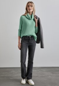 Mintgrön ribbad tröja med cowl-krage, svarta jeans och vita sneakers. Modell som håller en grå kappa. Enkel, avslappnad outfit.