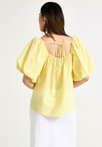 Gul texturerad blus med puffärmar och öppen rygg, med knytning. Lätt tyg med ett mjukt och luftigt utseende.