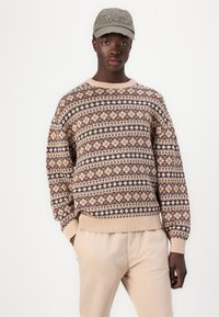 Strikket sweater med geometriske mønstre i beige, brun og mørkeblå. Rund hals, ribbede manchetter og kant; bæres over cremede bukser.