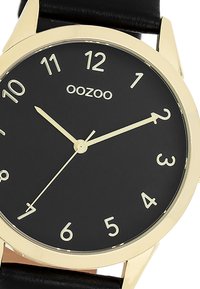 Montre à cadran noir avec des index et aiguilles dorés. Bracelet en cuir noir lisse, boîtier en métal rond, arborant le nom de la marque "oozoo."