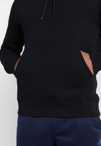 Felpa nera con cappuccio realizzata in tessuto morbido, dotata di tasca a marsupio frontale, cappuccio con cordino e polsini e orlo a costine; indossata sopra pantaloni blu scuro.