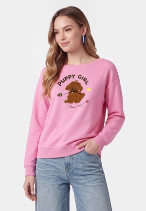 Ung kvinde med langt hår iført en lyserød sweatshirt med en brun hvalp og teksten "Puppy Girl I Love Puppies," kombineret med lyseblå jeans.