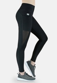 Schwarze Leggings mit hohem Bund, ausgestattet mit einem Mesh-Einsatz. Hergestellt aus einem dehnbaren Stoff, kombiniert mit grauen Sportschuhen.
