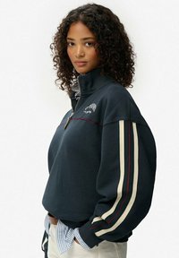 Pull en polaire bleu marine avec un col haut et une demi-zip, présentant des rayures crème et rouge sur les manches et un texte brodé sur la poitrine.