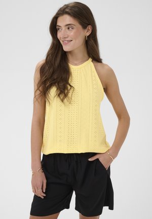 Mladá žena s dlhými hnedými vlasmi, ktorá má na sebe žltý tielko bez rukávov a čierne šortky, usmieva sa a pozerá do strany.