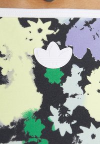 Bílé logo Adidas trefoil na barevném květinovém materiálu s abstraktními vzory květin v černé, zelené, fialové a žluté barvě.