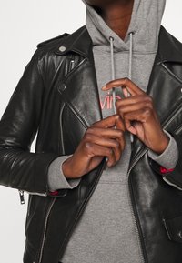 Giacca in pelle nera da motociclista con hardware argentato, tasche con zip, indossata sopra una felpa grigia con cappucci e dettagli rossi.