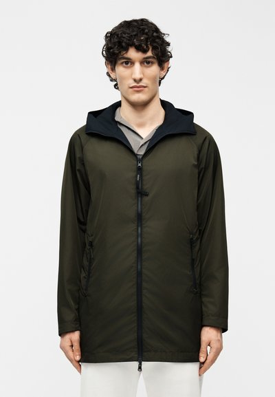 Homme aux cheveux bouclés et foncés portant une veste à capuche vert foncé avec une fermeture éclair noire sur une chemise grise et un pantalon blanc, debout droit.