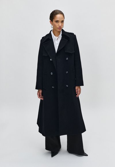 Cappotto doppiopetto nero con ampi rever, dettagli con bottoni e tasche laterali, realizzato in un tessuto spesso, abbinato a pantaloni neri e scarpe a punta.