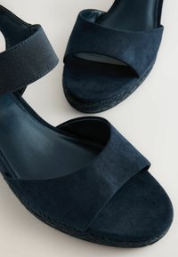Mørkeblå sandaler med en blød ruskindsoverflade, bred stropdesign og en flettet jute-sål. Har en justerbar stofankelrem.