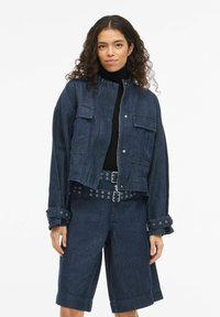 Tumša džinsa jaka ar īsu dizainu, aprīkota ar divām priekšējām kabatām un rāvējslēdzēja aizdari, ko papildina tumši džinsa šorti un josta ar metāla acīm.