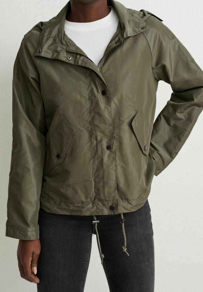 Personne portant une veste à capuche vert olive avec poches avant, cordons au bas et un t-shirt blanc côtelé en dessous.