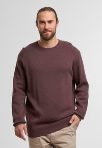 Weinroter strukturierter Strickpullover mit rundem Ausschnitt, gerippten Bündchen und Saum, getragen über hellen Hosen.