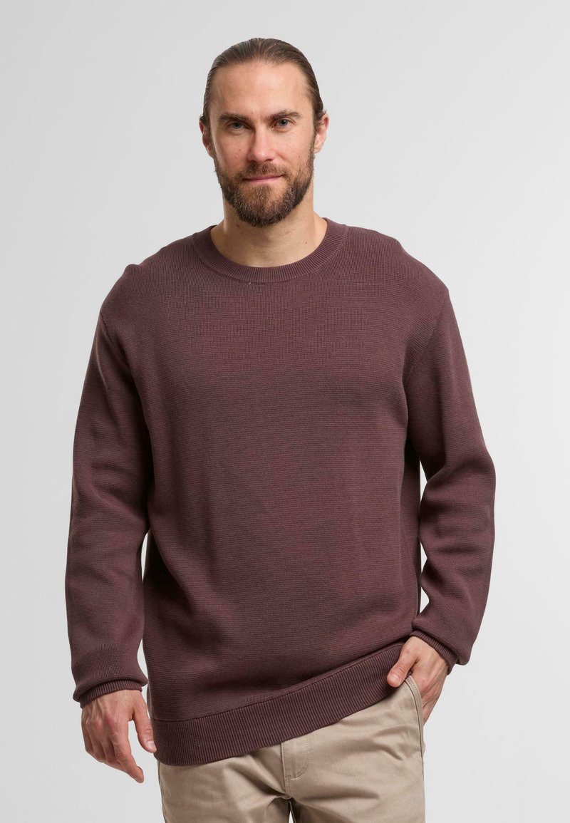 Weinroter strukturierter Strickpullover mit rundem Ausschnitt, gerippten Bündchen und Saum, getragen über hellen Hosen.