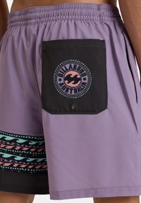 Violetinės maudymosi šortai su juodu kišeniu, kuriame yra apvalus logotipas. Dekoratyvinis bangų raštas teal ir rožinės spalvos palei apatinį kraštą. Minkšta tekstūra.