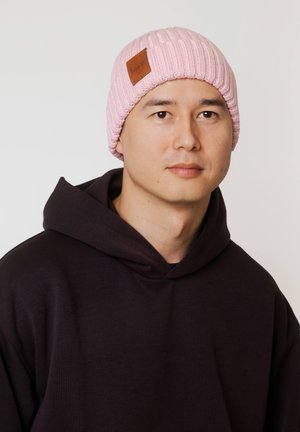 Różowa dziergana czapka typu beanie z prążkowaną teksturą i brązową skórzaną metką, noszona przez osobę w ciemnej bluzie z kapturem. Miękki materiał i dopasowany krój.