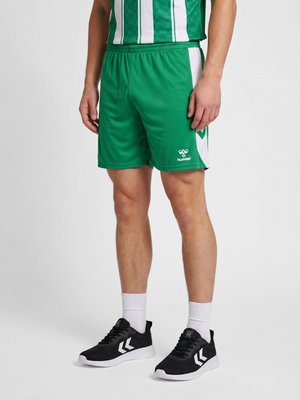 Mann trägt grüne und weiße Sportshorts, weiße Socken und schwarze Sportschuhe, steht vor neutralem Hintergrund.