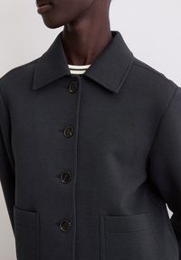 Ciemnoszary płaszcz wykonany z miękkiej tkaniny, z kołnierzem, z pięcioma guzikami z przodu i dwoma naszywanymi kieszeniami. Widoczny jest teksturowany materiał.