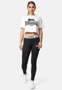 Black leggings with a Lonsdale logo, paired with a white cropped t-shirt featuring a lion graphic. Model wears white sneakers.
